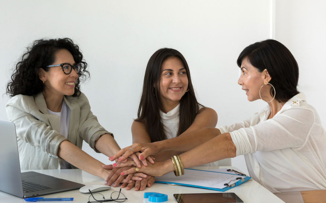 Tres mujeres reunidas, en una mesa de trabajo, juntan sus manos y comparten miradas de complicidad, para ejemplificar la importancia del liderazgo femenino