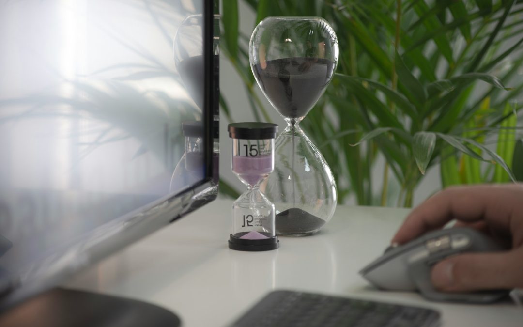 Hombre trabaja en computadora, sosteniendo el mouse, con dos relojes de arena activos en el fondo.