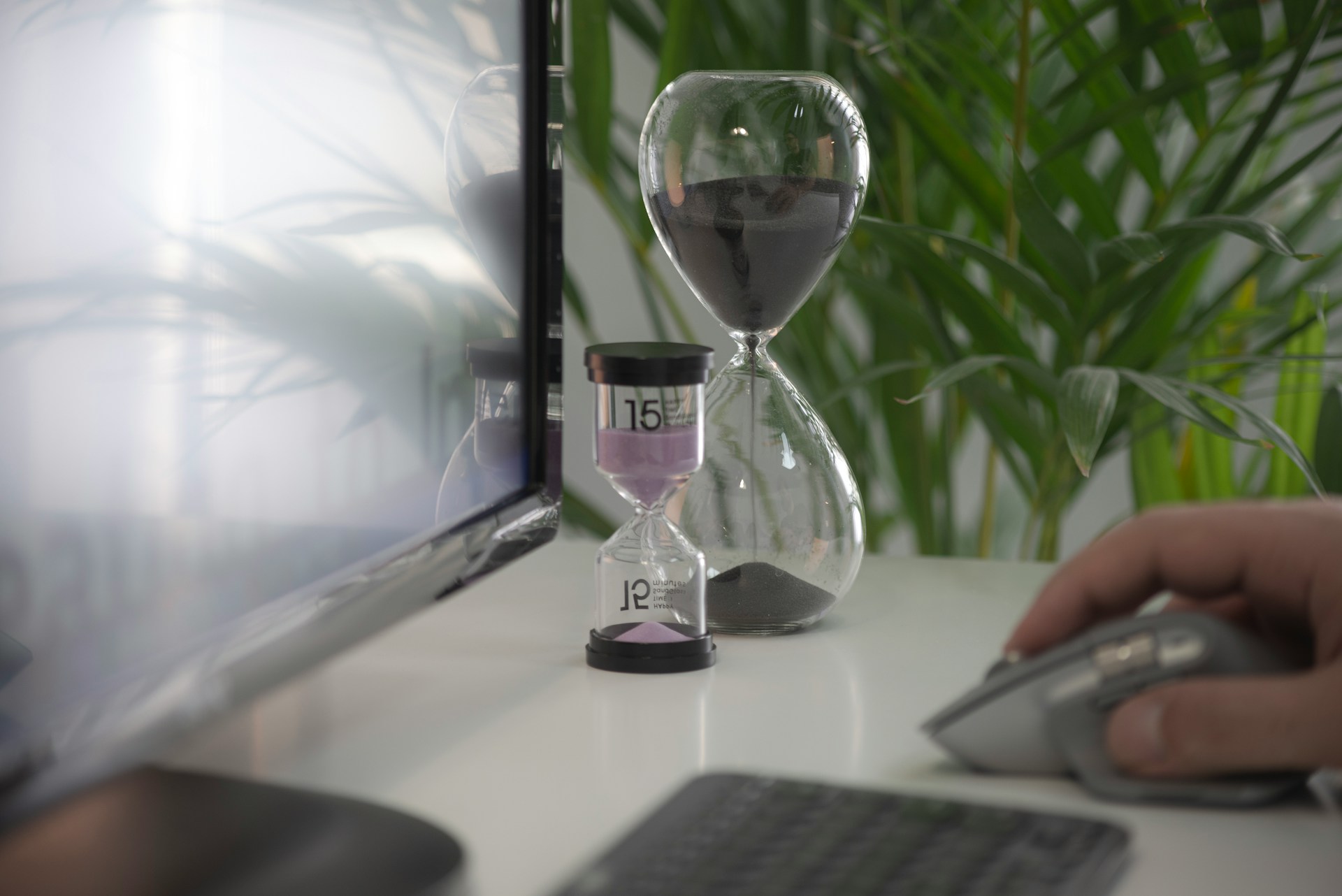 Hombre trabaja en computadora, sosteniendo el mouse, con dos relojes de arena activos en el fondo.