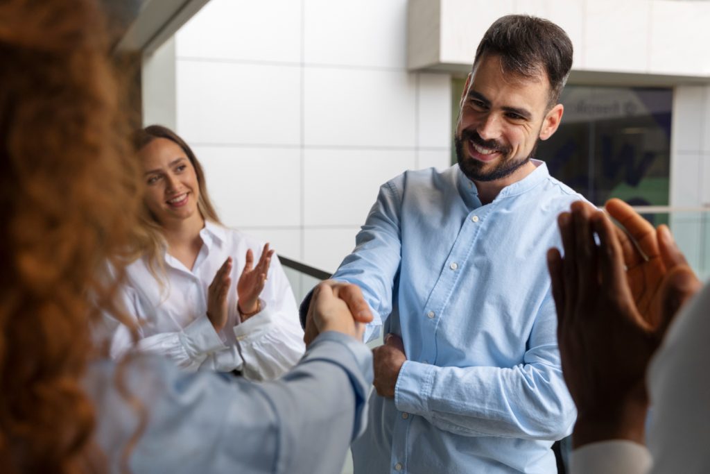 Reunión de trabajo con un hombre estrechando la mano de un colaborador, al momento que delega actividades, mientras un equipo diverso sonríe y aplaude. 