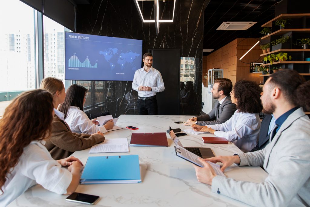 Un hombre presenta proyecciones de un negocio, en una pantalla, frente a un grupo de inversionistas en una sala de reuniones moderna, ilustrando los beneficios de un plan de negocios. 