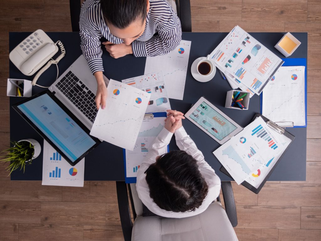 Dos personas en un escritorio revisan gráficos, en la mesa se encuentra una computadora y una tableta que muestran herramientas digitales en sus pantallas.
