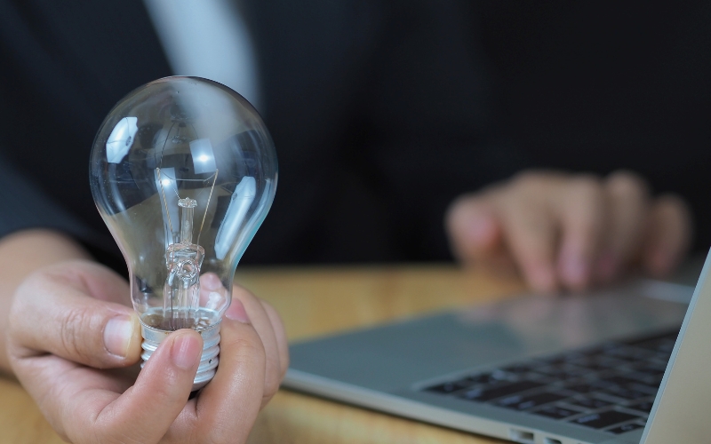 Persona sostiene foco junto a laptop como símbolo de innovación en un proyecto de emprendimiento de alto impacto.