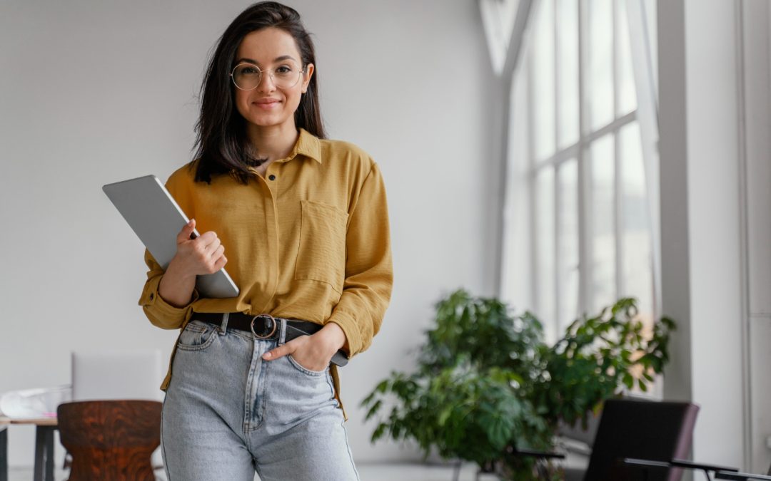 Mujer joven con ropa casual y lentes mira hacia la cámara. Con una mano sostiene una tableta. La imagen representa a una mujer emprendedora.