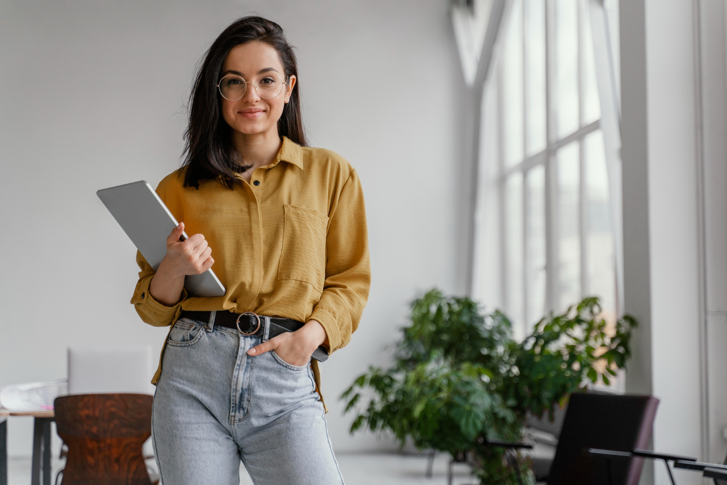 Mujer joven con ropa casual y lentes mira hacia la cámara. Con una mano sostiene una tableta. La imagen representa a una mujer emprendedora.
