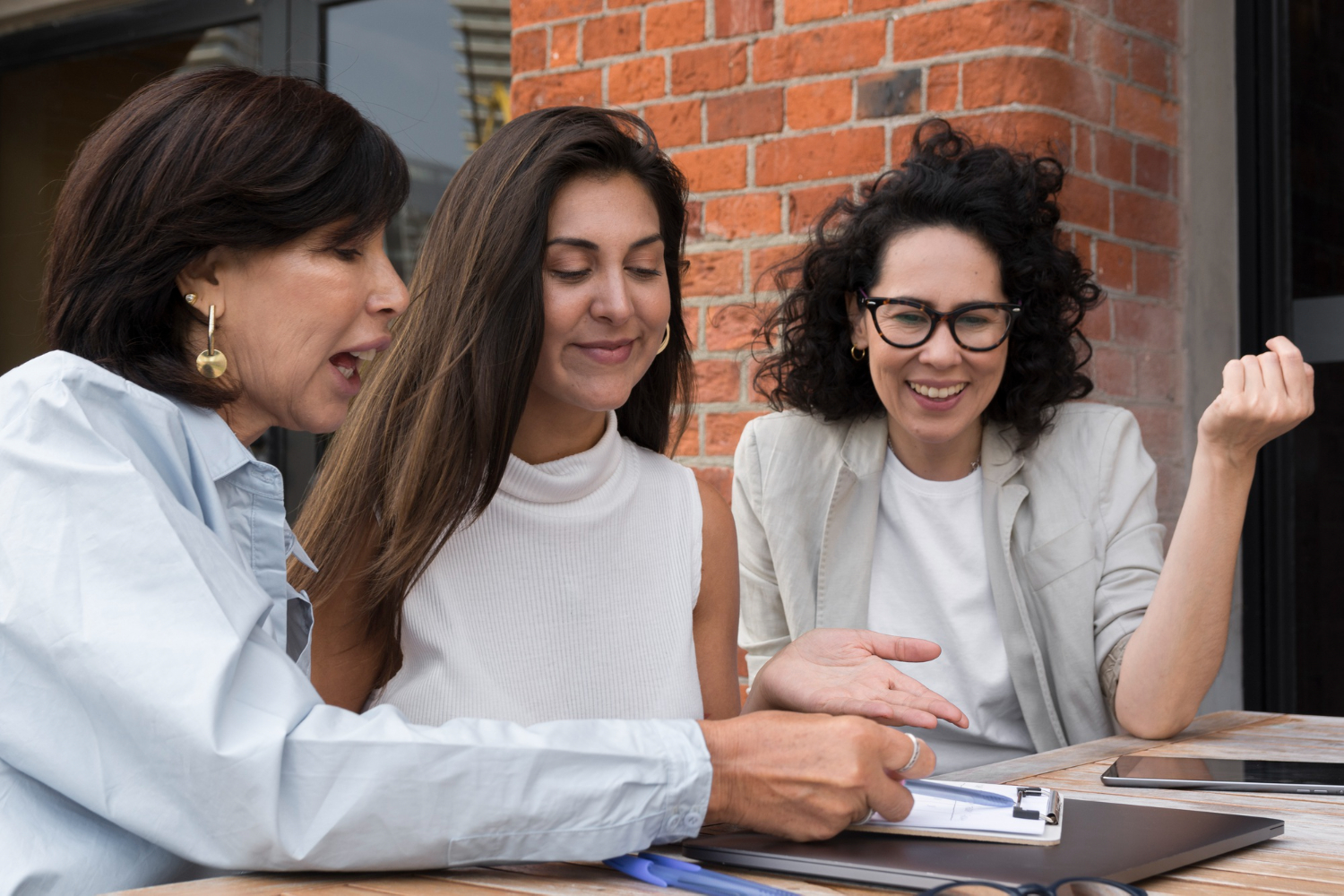 Tres mujeres reunidas en una mesa al aire libre. La mujer de mayor edad muestra datos, que se encuentran en un documento, a las mujeres más jóvenes que ponen atención.