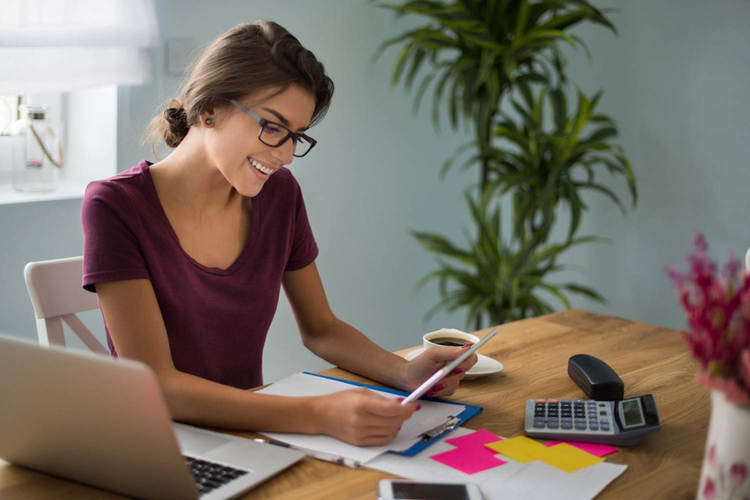  Mujer sentada revisa información en una tableta electrónica. En su escritorio hay una computadora, una calculadora, documentos y una taza con café.