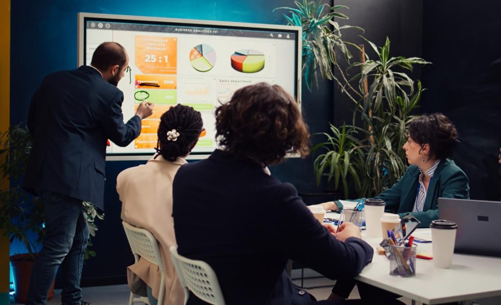 Un hombre encabeza una presentación ante un grupo de personas, en una sala de juntas. En una pantalla muestra una serie de gráficas.